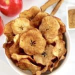Air Fryer Apple Chips in white bowl with apples and cinnamon sticks in background