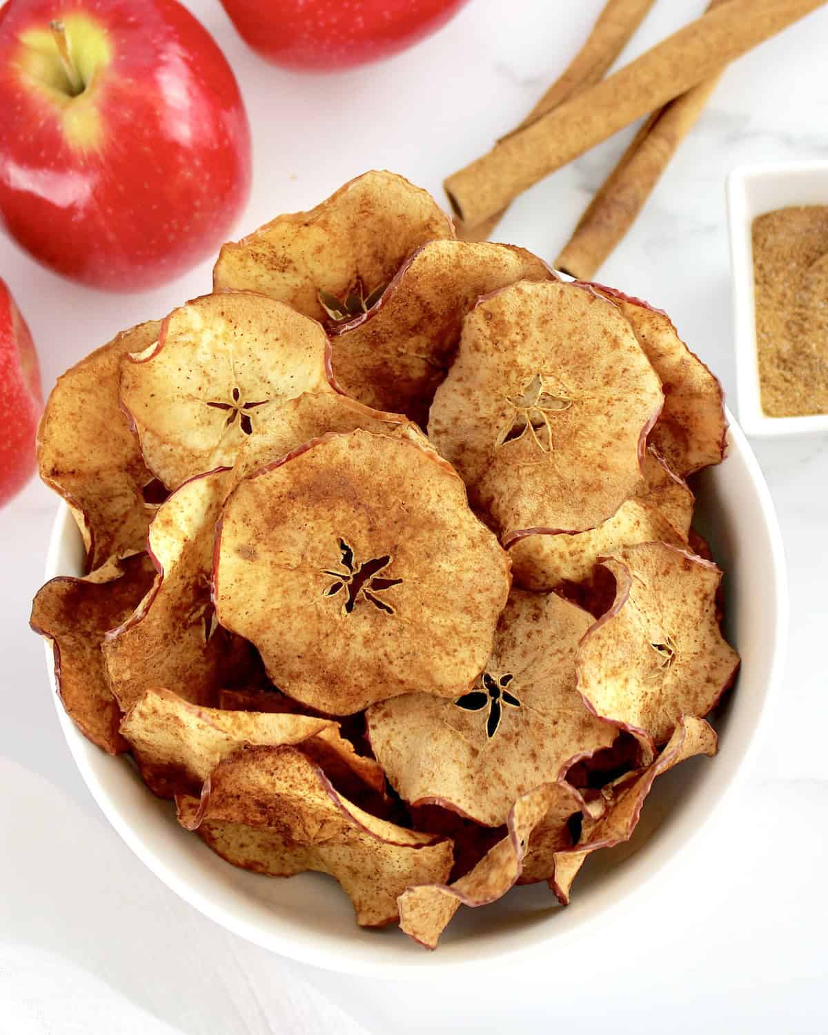 Air Fryer Apple Chips in white bowl with apples and cinnamon sticks in background