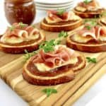 Baked Brie and Prosciutto Crostini on cutting board with apple butter jar and round white plates in background