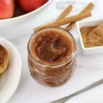 Slow Cooker Apple Butter in open glass jar with apples in background with cinnamon sticks and toast