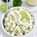 Easy Coconut Rice in white bowl with spoon, chopped cilantro and 2 small lime wedges on top with limes in back