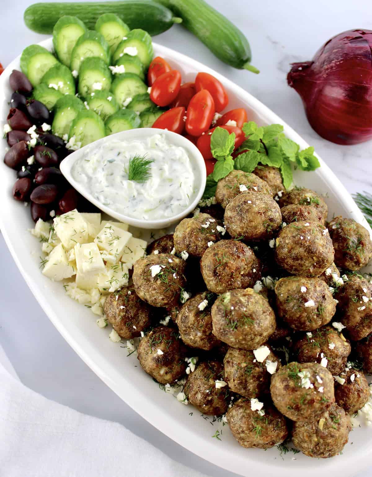 Greek Meatballs on white platter with cucumbers, tomatoes, olives and dip