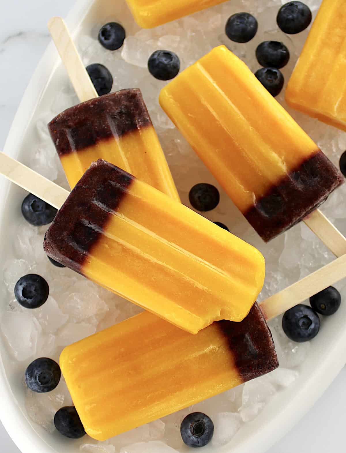 Mango Blueberry Popsicles on white oval platter with crushed ice and fresh blueberries around