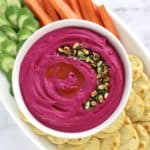 Roasted Beet Hummus in white bowl with crackers, carrot sticks and cucumber slices on white plate