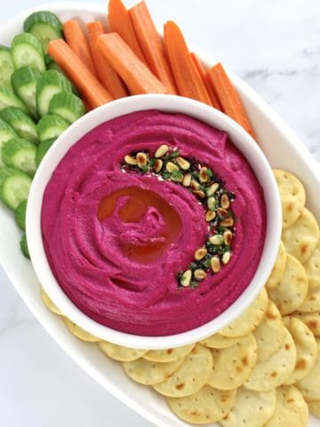 Roasted Beet Hummus in white bowl with crackers, carrot sticks and cucumber slices on white plate