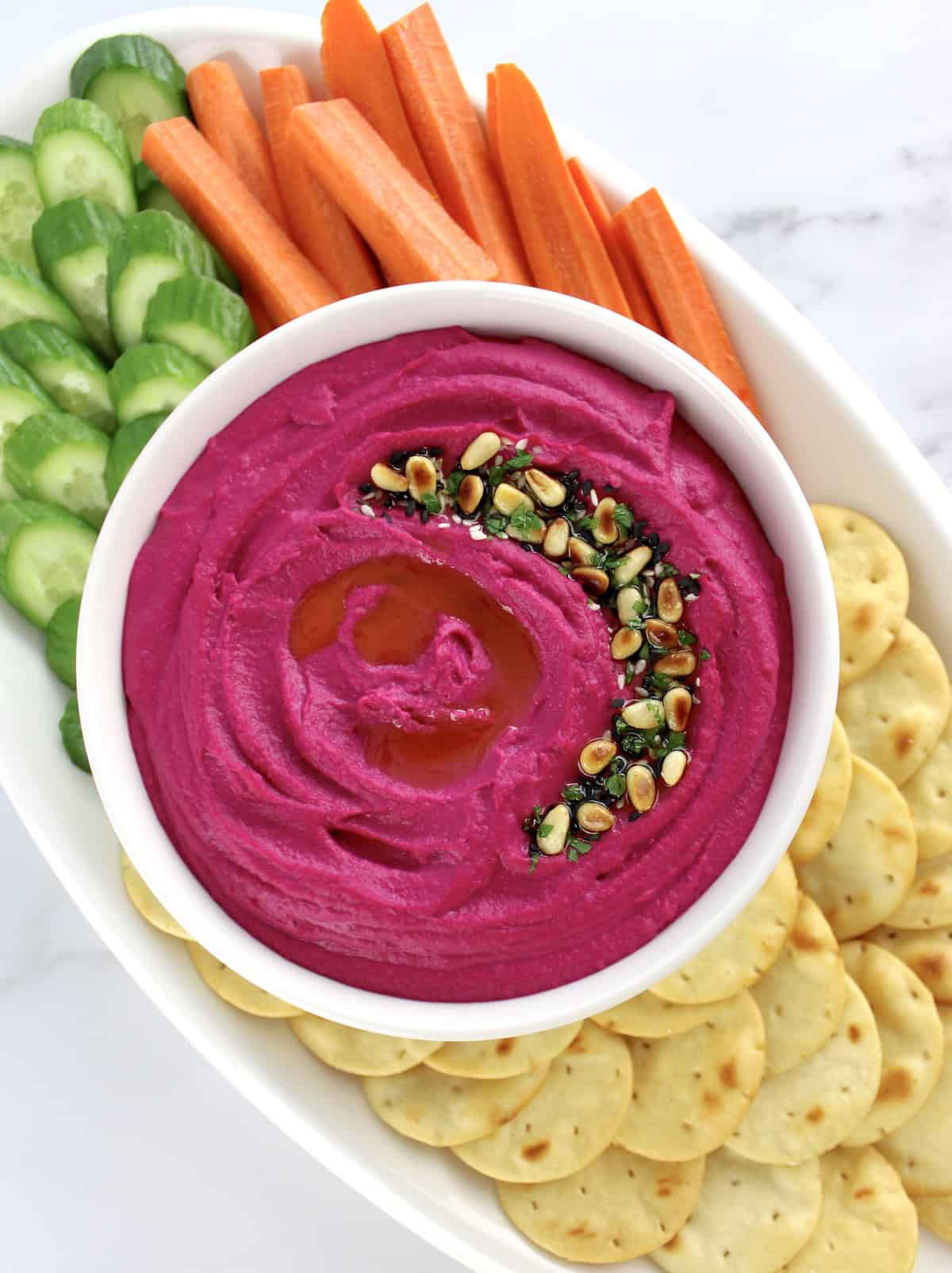 Roasted Beet Hummus in white bowl with crackers, carrot sticks and cucumber slices on white plate