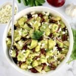 overhead view of Greek Potato Salad in white bowl with silver serving spoon and veggies in back