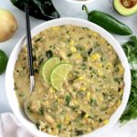 White Chicken Chili in white bowl with 2 lime slices and spoon in bowl with veggies in back