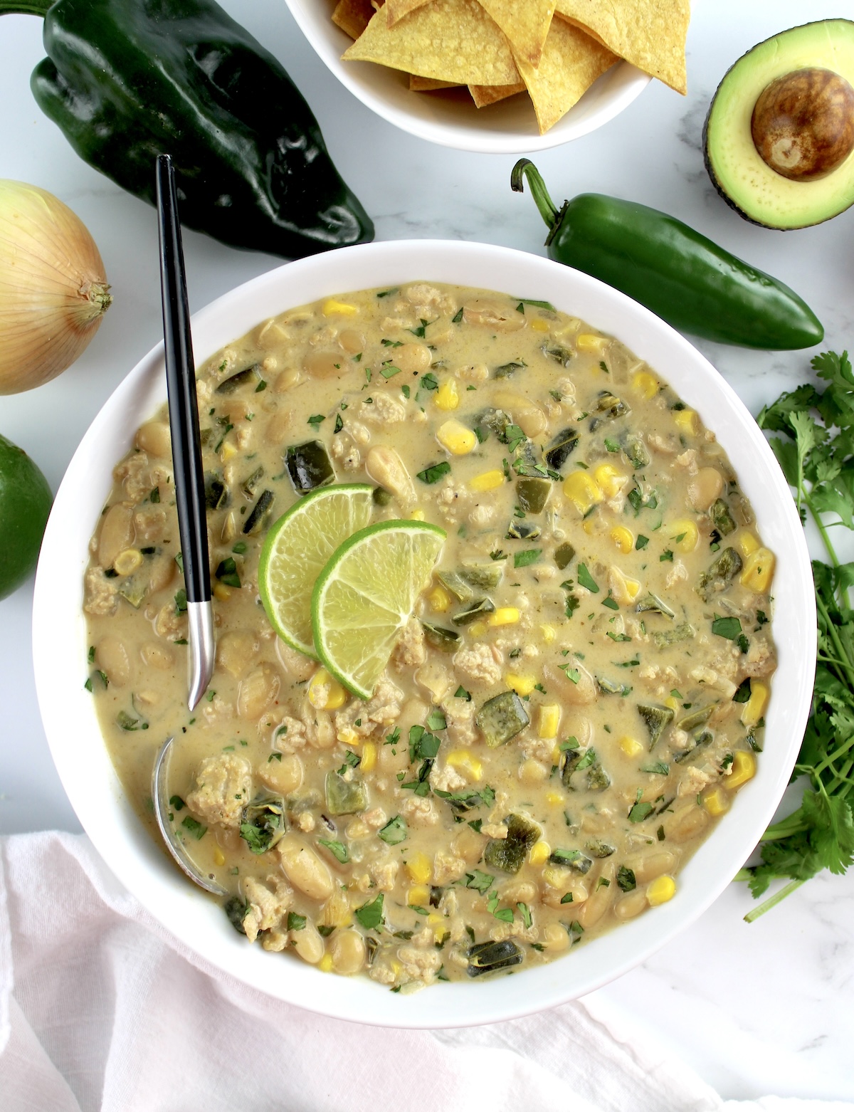 White Chicken Chili in white bowl with 2 lime slices and spoon in bowl with veggies in back