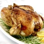 whole roasted chicken on white platter with fresh rosemary and lemon slices