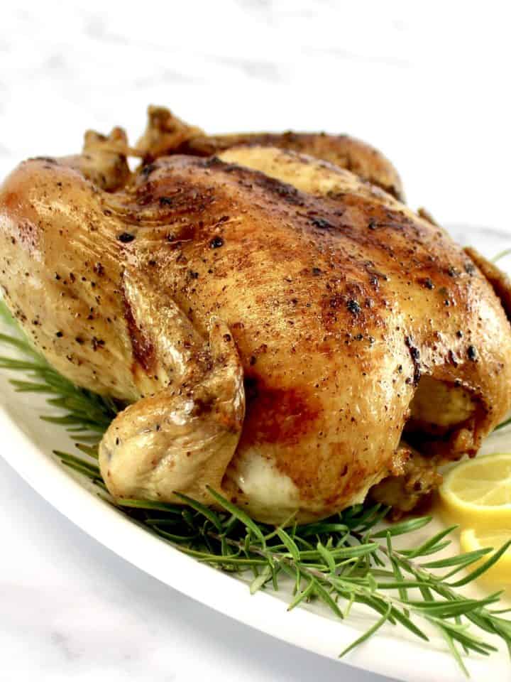 whole roasted chicken on white platter with fresh rosemary and lemon slices