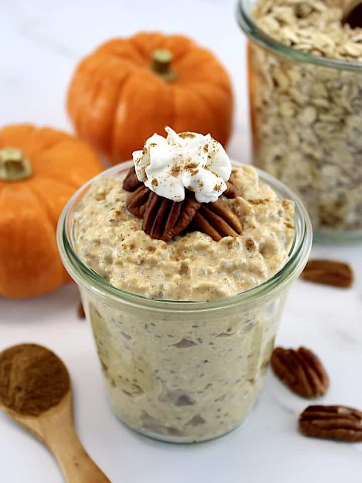 Pumpkin Pie Overnight Oats in open glass jar with whip cream on top and pumpkins in back