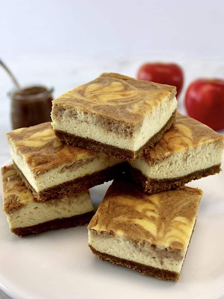 Apple Butter Cheesecake Bars stacked up on white platter with apples and apple butter in back