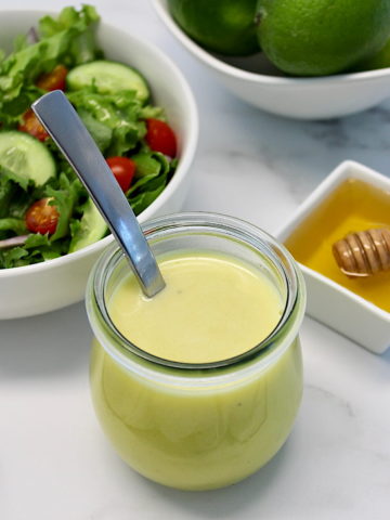 Honey Lime Vinaigrette in open glass jar with spoon in it, salad, honey and lime in back