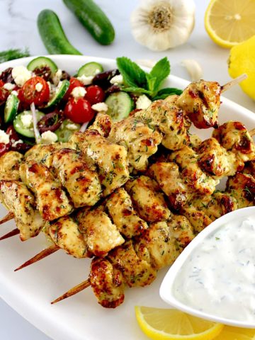 Air Fryer Greek Chicken Souvlaki on white platter with tzatzi sauce and Greek salad on side