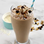 Almond Joy Protein Shake in glass with whip cream, chocolate chips and shredded coconut on top with blue straw