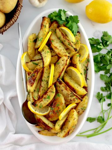 Greek Lemon Potatoes on white oval platter with lemon slices and parsley with serving spoon on side